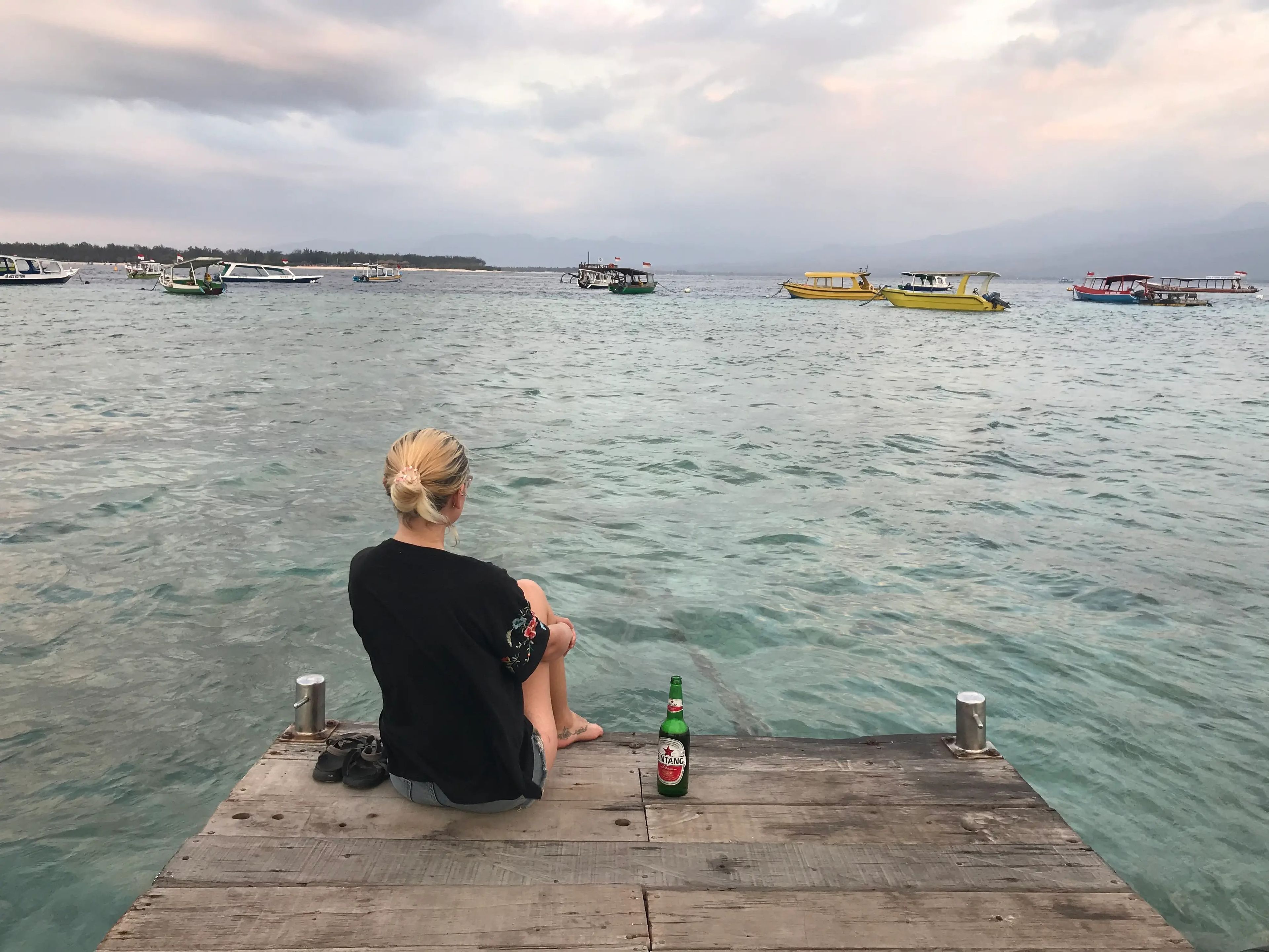 Jennifer Sizeland sitting on the edge of a dock looking at the water drinking a beer.